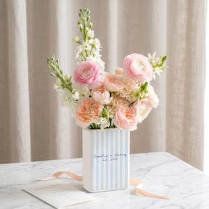 May include: A rectangular ceramic vase with light blue vertical stripes, featuring the text Amelia + Harry est. 2024. The vase is filled with a bouquet of pink and peach ranunculus flowers and white snapdragons, accompanied by a printed card.