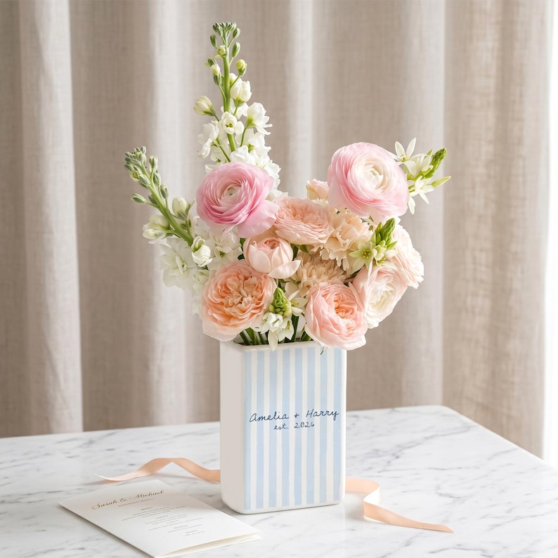 May include: A rectangular ceramic vase with light blue vertical stripes, featuring the text Amelia + Harry est. 2024. The vase is filled with a bouquet of pink and peach ranunculus flowers and white snapdragons, accompanied by a printed card.
