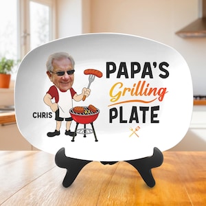May include: White oval serving plate with the text "PAPA'S Grilling PLATE" in black and orange. The plate features a cartoon illustration of a man grilling, with the name "CHRIS" on the left side. The plate is displayed on a black stand.