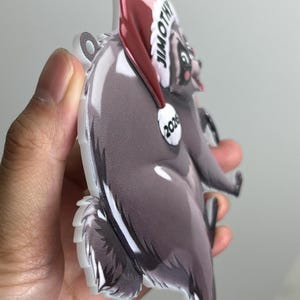 Peut inclure: Une d&eacute;coration en acrylique plate et d&eacute;coup&eacute;e en forme de raton laveur portant un bonnet de P&egrave;re No&euml;l rouge. Le bonnet porte l'inscription JIMOTHY et l'ann&eacute;e 2026 en blanc. Le raton laveur a un pelage gris, un ventre blanc et un trou.