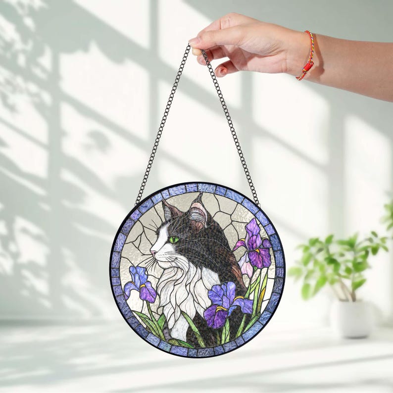 Peut inclure: Panneau de vitrail rond repr&eacute;sentant un chat noir et blanc aux yeux verts, entour&eacute; d'iris violets. L'&oelig;uvre est encadr&eacute;e d'une bordure bleue et suspendue par une cha&icirc;ne.