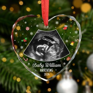 May include: Heart-shaped clear glass Christmas ornament with a red ribbon. The ornament features a sonogram image and the text "Baby William ARRIVING 2026" with festive holiday graphics. The ornament is hanging in front of a blurred background.