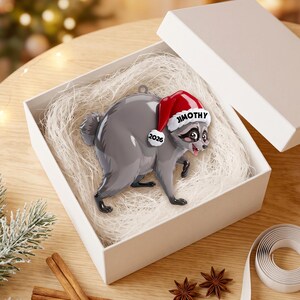 Peut inclure: Une d&eacute;coration en forme de raton laveur gris portant un bonnet de P&egrave;re No&euml;l rouge et blanc avec le nom JIMOTHY imprim&eacute; sur le bord et l'ann&eacute;e 2026 sur le c&ocirc;t&eacute;. Le raton laveur a des taches noires autour des yeux et une petite queue.