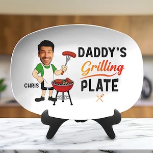 May include: White oval plate featuring a cartoon of a man grilling, with the text "DADDY'S Grilling PLATE" and the name "CHRIS". A small grill graphic is also present. The plate is displayed on a black stand.