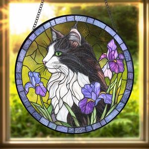Peut inclure: Vitrail rond repr&eacute;sentant un chat noir et blanc aux yeux verts, entour&eacute; d'iris violets et de feuillage vert. La fen&ecirc;tre a une bordure bleue et violette et est suspendue par une cha&icirc;ne. Le fond est jaune et vert clair.