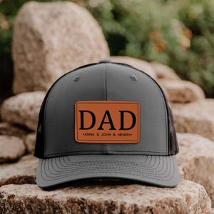 May include: Grey trucker cap with a black mesh back and a brown leather patch. The patch displays the word "DAD" in bold black letters, with "ANNA & JOHN & HENRY" underneath.