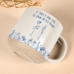 May include: Cream ceramic mug with a speckled handle and a rough, unglazed base. The mug features blue floral illustrations and the text "IT TAKES A LONG TIME TO GROW AN OLD FRIEND" with the names "Diana" and "Anna" below the flowers.