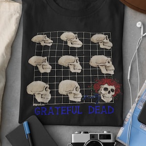 May include: Black t-shirt featuring a grid of ape skulls and a floral skull design. The text "Grateful Dead" is printed in blue below the design. The shirt is surrounded by accessories including a hat, sunglasses, and a camera.