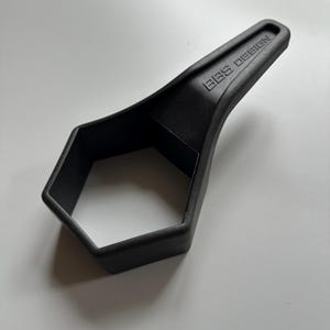 Puede incluir: Una herramienta negra de forma hexagonal con un mango que lleva el texto "BBS DESIGN". Fabricada en material negro mate, esta herramienta parece ser una llave especial o un dispositivo similar, probablemente para un uso específico.