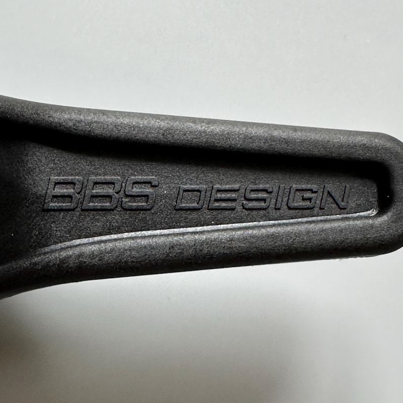 Pu&ograve; includere: Primo piano di un oggetto grigio scuro con la scritta in rilievo "BBS DESIGN". L'oggetto ha una superficie leggermente strutturata e sembra essere un componente o parte di un elemento pi&ugrave; grande. L'illuminazione evidenzia le lettere in rilievo.