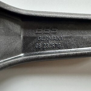 Pu&ograve; includere: Primo piano di un componente automobilistico grigio scuro con il testo "BBS GERMANY 09.23.576" in rilievo sulla superficie. Il componente ha un design curvo ed ergonomico.