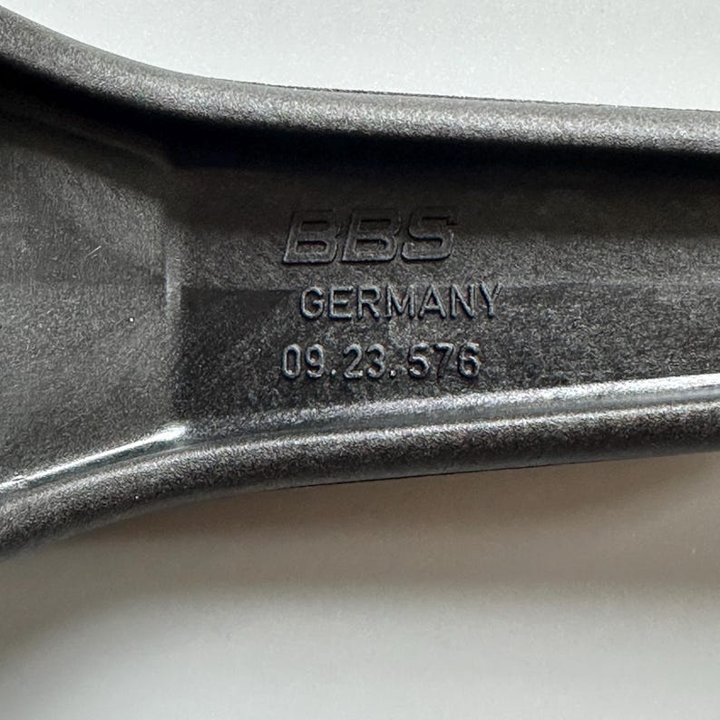 Pu&ograve; includere: Primo piano di un componente automobilistico grigio scuro con il testo "BBS GERMANY 09.23.576" in rilievo sulla superficie. Il componente ha un design curvo ed ergonomico.