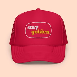 May include: Red trucker hat with a white oval patch that says "stay golden" in white and yellow lettering.