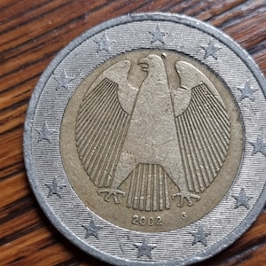 Puede incluir: Una moneda de 2 euros alemana de 2002 de color plateado y dorado con una representación del águila alemana en el anverso. La moneda tiene 12 estrellas alrededor del borde.