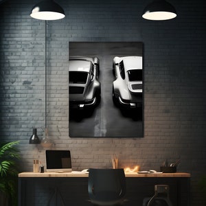 May include: A framed canvas print featuring two classic sports cars, one silver and one white, viewed from the rear. The artwork hangs above a desk with a laptop, chair, and desk accessories. The room has a modern, industrial aesthetic with exposed brick and pendant lighting.