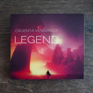May include: Square CD case with a fantasy landscape design. The cover features the text "CRUENTA VENGANZA" above the word "LEGEND" in white. The artwork depicts a lone figure walking towards a futuristic city with a red and purple sky.