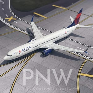Pode incluir: Um modelo de avião Delta Airlines branco e azul com detalhes vermelhos na cauda, ​​repousando sobre uma superfície cinza com marcações amarelas e brancas. O texto "PNW" e "PACIFIC NORTHWEST TAXIWAY" são visíveis.