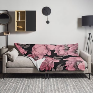 May include: A pink and black floral throw blanket with a soft, plush texture. The blanket features a repeating pattern of large pink peonies on a black background.