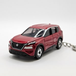 May include: A red toy car keychain of a Nissan X-Trail SUV. The car has a black grille and headlights, and a silver roof rack.