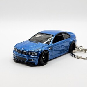 May include: A blue die-cast model car with black wheels and a silver keychain ring. The car has a metallic finish and detailed features, including a front grill and headlights. The car is a coupe style.