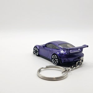May include: A purple die-cast model car keychain. The car has a spoiler and black and white wheels. The keychain is made of silver metal and has a split ring. The car is a miniature replica of a sports car.