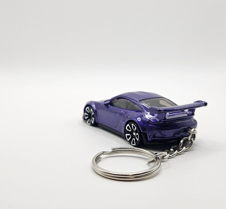 May include: A purple die-cast model car keychain. The car has a spoiler and black and white wheels. The keychain is made of silver metal and has a split ring. The car is a miniature replica of a sports car.