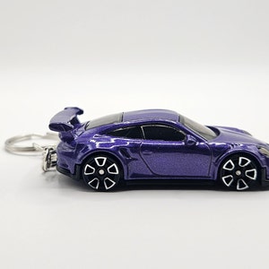 May include: A purple die-cast model car keychain. The car has black and white wheels and a rear spoiler. The keychain ring is silver. The car is a miniature replica of a sports car.