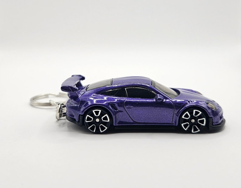 May include: A purple die-cast model car keychain. The car has black and white wheels and a rear spoiler. The keychain ring is silver. The car is a miniature replica of a sports car.