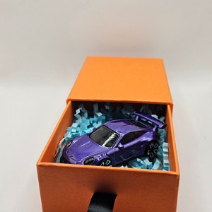May include: A purple toy sports car with a rear wing sits inside an orange gift box filled with blue paper shreds. The box has a black ribbon pull and is set against a white background.