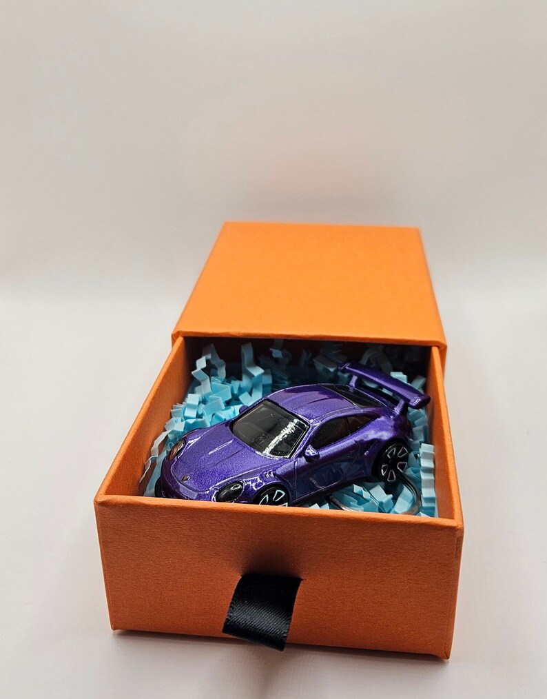 May include: A purple toy sports car with a rear wing sits inside an orange gift box filled with blue paper shreds. The box has a black ribbon pull and is set against a white background.