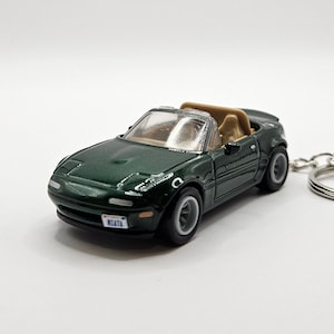 May include: A dark green miniature convertible car with a tan interior and a license plate that reads "MIATA". The toy car has a silver keychain attached. The car is a detailed replica of a classic roadster.