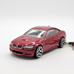 May include: A red miniature car model of a BMW 3 Series Coupe, with a silver grille and black wheels. The car is attached to a silver key chain.