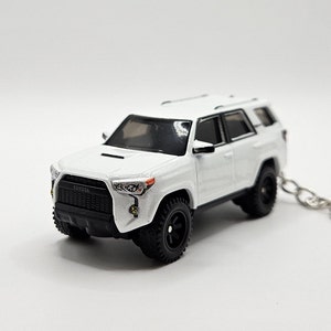 May include: White Toyota 4Runner key chain. The keychain is a miniature replica of the SUV with black wheels and a black front grille. The word "TOYOTA" is visible on the front grille.