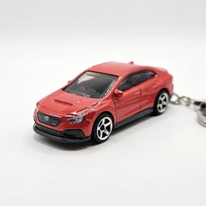 May include: A red die-cast toy car keychain featuring a sedan model with a hood scoop, black front grille, clear windows, and silver five-spoke wheels. The vehicle is attached to a standard metal split ring for keys, showcasing a detailed exterior.