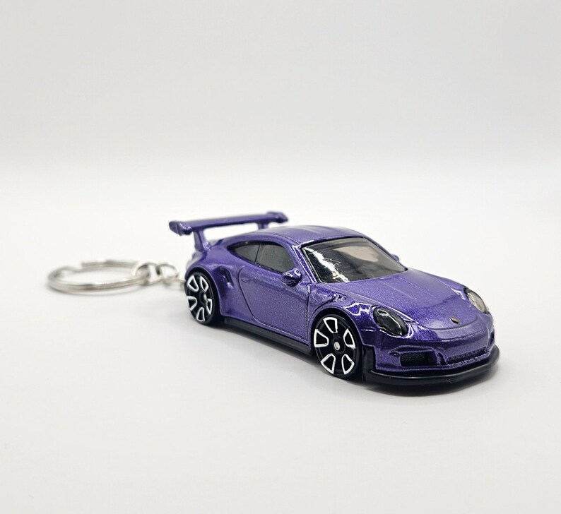 May include: A purple die-cast model car keychain. The miniature sports car features a rear spoiler, black and white wheels, and a silver key ring. The car has a glossy finish and is set against a white background.