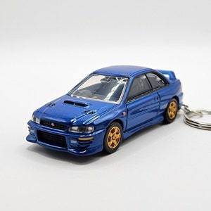 May include: A blue die-cast model car with gold-colored wheels and a keychain attachment. The car has a spoiler and detailed features, showcasing a sporty design. The model is a miniature replica of a sports car.