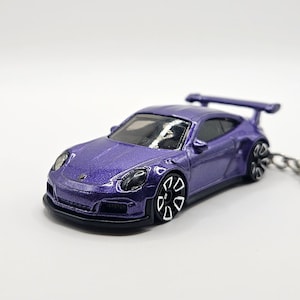 May include: A purple die-cast model car with a rear spoiler and black accents. The toy car is attached to a keychain. The car has black wheels with white rims and a black front bumper.