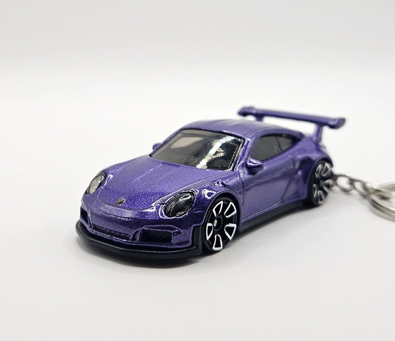 May include: A purple die-cast model car with a rear spoiler and black accents. The toy car is attached to a keychain. The car has black wheels with white rims and a black front bumper.