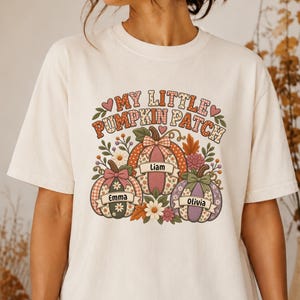 May include: An ivory short-sleeve t-shirt featuring a graphic print with the text "MY LITTLE PUMPKIN PATCH" above three illustrated pumpkins. Each pumpkin displays a name: "Emma," "Liam," and "Olivia," decorated with floral and plaid patterns.