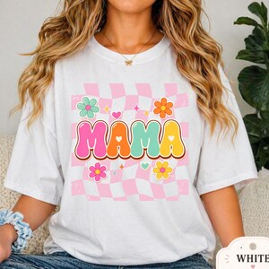 Puede incluir: Camiseta blanca con un diseño retro con la palabra "MAMA" en letras de burbujas coloridas. El diseño incluye un fondo a cuadros rosa y blanco, flores y corazones. La camiseta es de corte holgado.