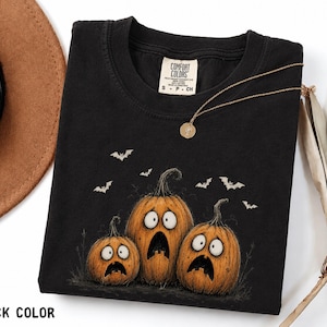 May include: A black short-sleeve t-shirt featuring a graphic of three orange pumpkins with wide, shocked expressions and black outlines. Small white bats are flying above the pumpkins. The shirt has a Comfort Colors brand label at the neck.