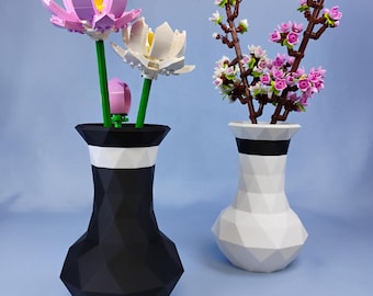 Puede incluir: Dos jarrones decorativos, uno negro y otro blanco, cada uno con flores artificiales. El jarrón negro presenta un diseño geométrico con una banda blanca. El jarrón blanco tiene un diseño similar. Las flores incluyen flores de loto rosas y blancas y flores rosas.