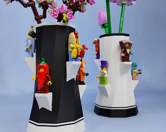 Puede incluir: Dos jarrones decorativos, uno negro y otro blanco, cada uno con un diseño geométrico único. Los jarrones están adornados con figuras de Lego y contienen flores artificiales. El jarrón negro tiene flores rosas, y el jarrón blanco tiene flores blancas y rosas.