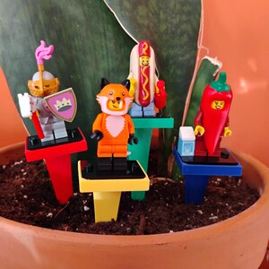 May include: A collection of Lego minifigures, including a knight, a fox, a hot dog, and a chili pepper, each standing on a colorful plant stake. The figures are set in a terracotta pot filled with soil, with a green plant in the background.