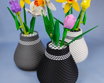 Puede incluir: Tres jarrones decorativos en negro, gris y blanco, cada uno con coloridas flores de Lego. Los jarrones tienen un diseño en espiral y una banda con estampado blanco y negro cerca de la parte superior. Las flores incluyen narcisos amarillos, narcisos blancos, tulipanes morados y tulipanes rosas.
