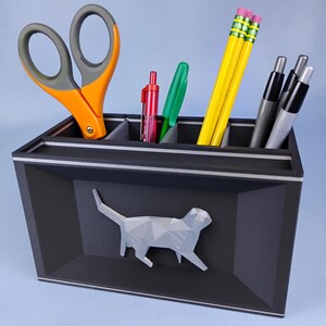 May include: A black desk organizer with a geometric cat design on the front. The organizer holds orange scissors, pens, pencils, and markers. The organizer has a gray and white trim.