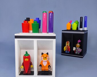 Peut inclure: Un organisateur de bureau blanc à deux compartiments contenant une figurine de piment et une figurine de renard, surmonté de marqueurs colorés. Un second organisateur noir se trouve derrière, avec un hot-dog et un chevalier en figurine.