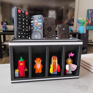 May include: A black and white remote control organizer with four compartments, each containing a Lego figurine. The top section holds four black remote controls. The figurines include a red chili pepper, an orange fox, a hot dog, and a knight.