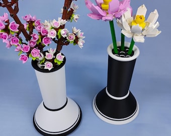 Puede incluir: Dos jarrones decorativos, uno blanco y otro negro, cada uno con arreglos florales. El jarrón blanco contiene una rama con flores rosas y blancas. El jarrón negro contiene dos flores, una rosa y otra blanca, con centros amarillos.