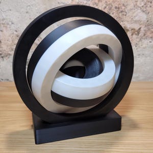 May include: A black and white abstract sculpture with a circular design. The sculpture is made of multiple interlocking rings and is mounted on a black base.